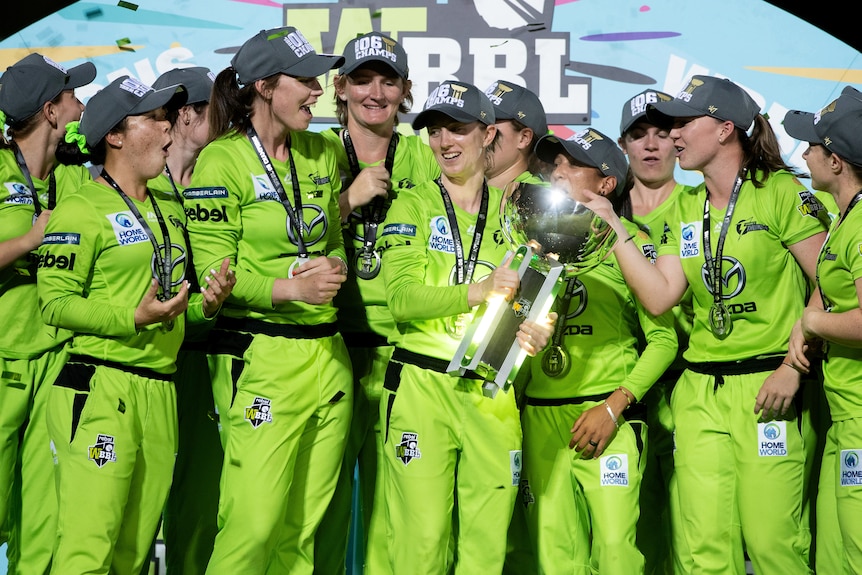Sydney Thunder players smiles and stand around a large silver trophy in front of a colourful backdrop