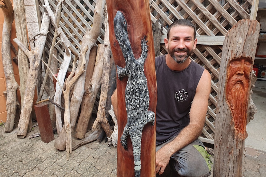 Man sitting between two tall logs he has carved, there is a face carved into one log and a goanna carved on the other