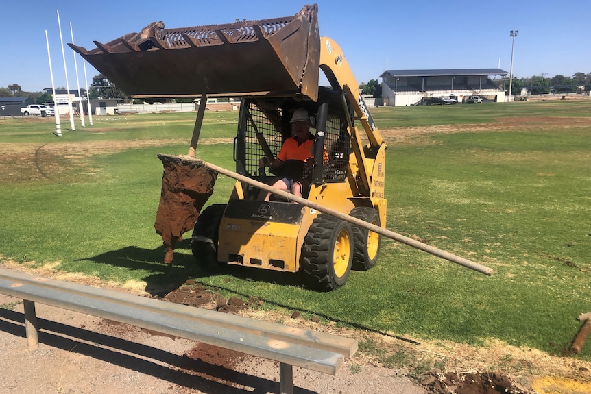A man in a bobcat pulling a fence post out of the ground on a football oval 