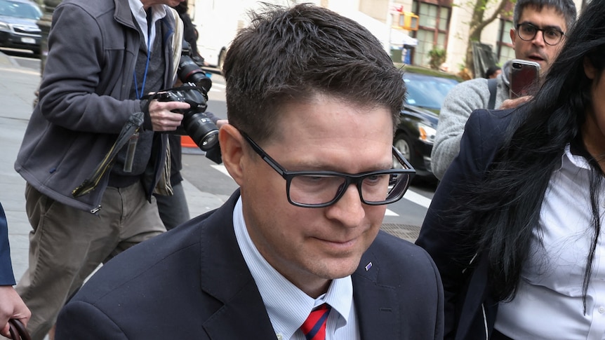 A suited man looks down as he leaves a courthouse in a wheelchair surrounded by media. 