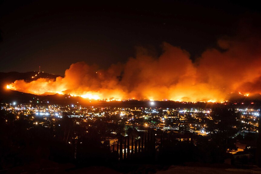 A valley at night, with hundreds of houses in it. Behind this a fire rages, smoke billowing into the air.
