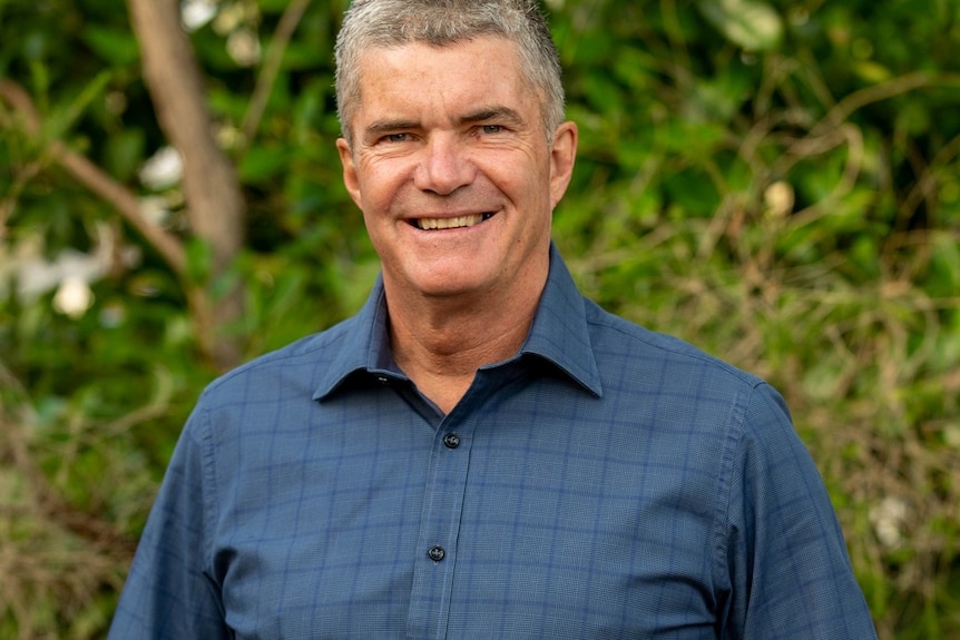 A middle-aged man with short, grey hair stands smiling front of some greenery.