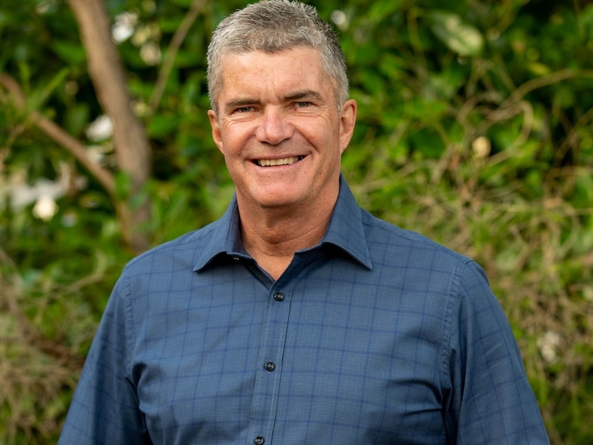 A middle-aged man with short, grey hair stands smiling front of some greenery.