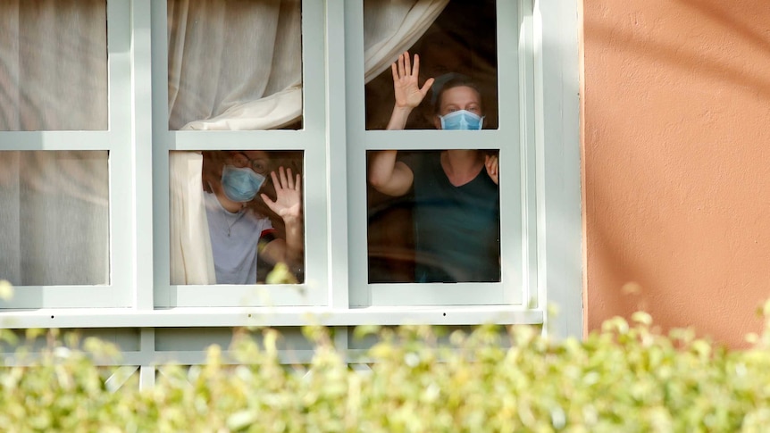 Two women wearing protective face masks, wave from the window of their hotel room.