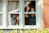 Two women wearing protective face masks, wave from the window of their hotel room.