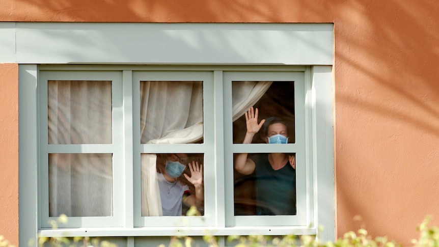 Two women wearing protective face masks, wave from the window of their hotel room.