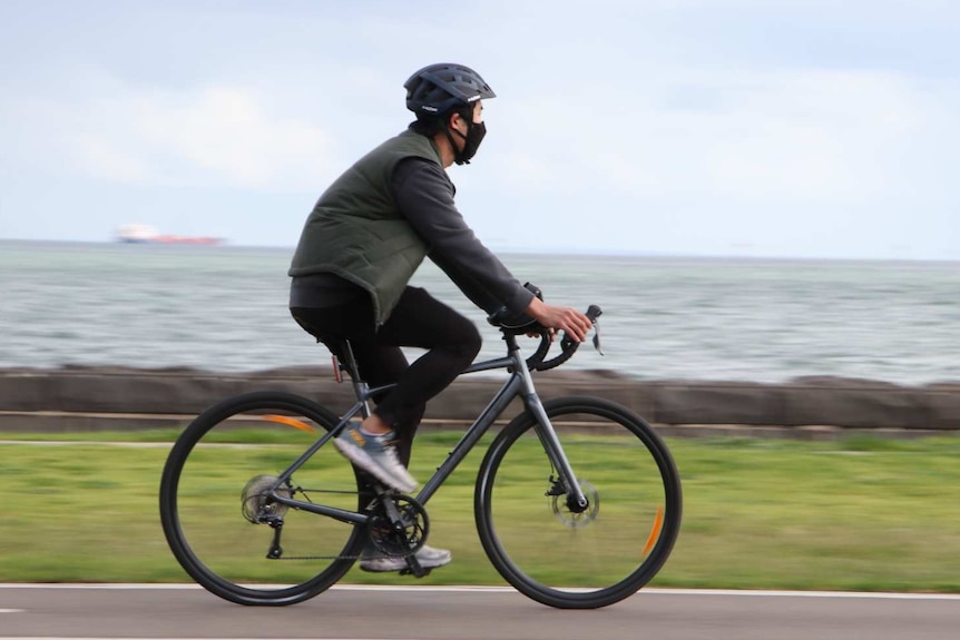 A man wearing a mask rides his bike beside water.