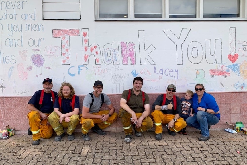Five men in firefighting trousers, a child and woman squat in front of a white wall with "Thank you" written in chalk behind the