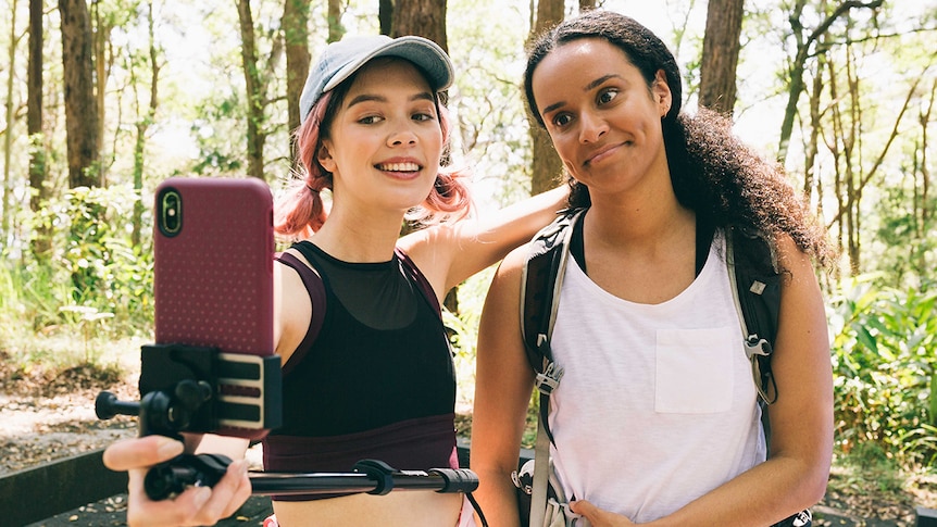 Two girls wearing active wear pose for a selfie during a hike on a sunny day in front of trees.