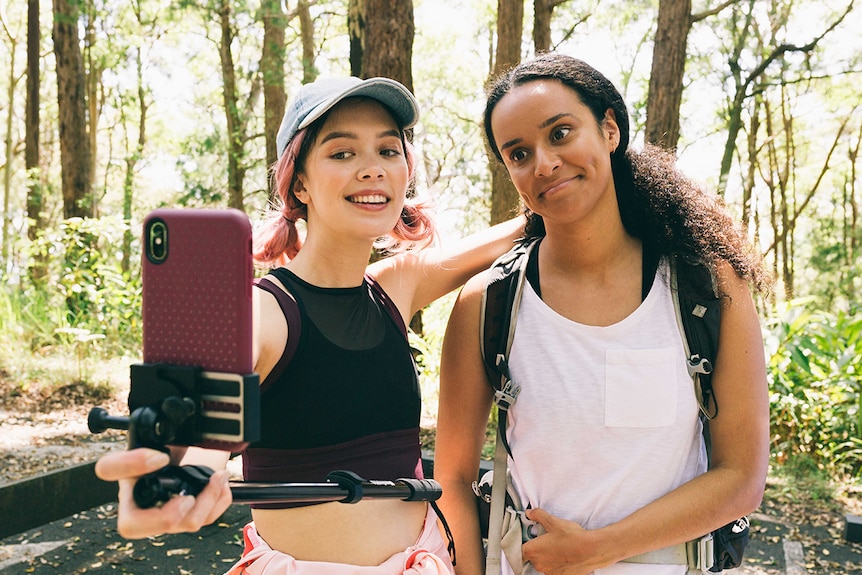 Two girls wearing active wear pose for a selfie during a hike on a sunny day in front of trees.