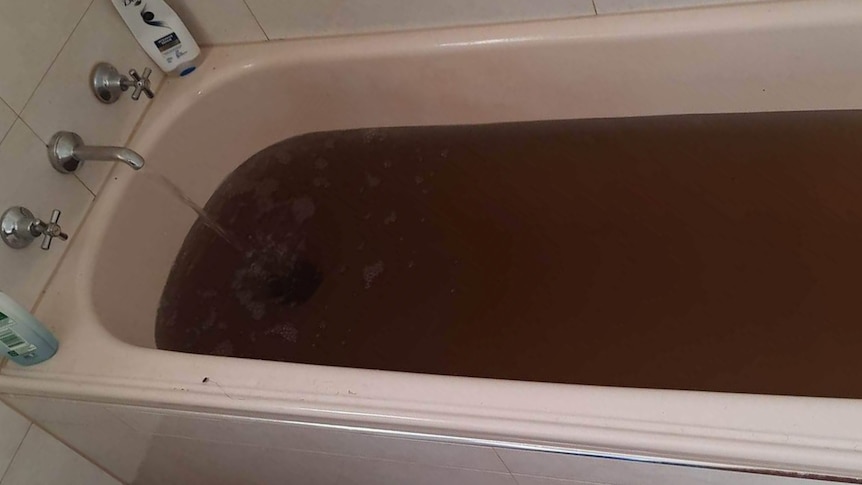 A pink bathtub filled with red-brown water. The tap is running and you can not see through the water. The tiles are white.