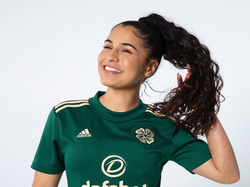 A woman in a green soccer shirt smiles and tosses her hair aside in front of a white background