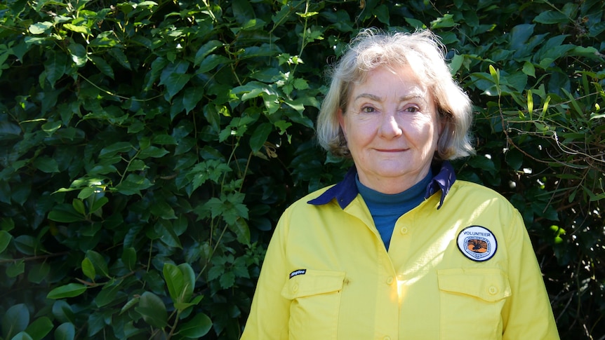 Sue Baker wearing a National Parks volunteer top smiling in front of lush green bush.