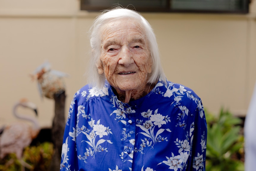 An elderly woman wearing a blue shirt with white flowers smiles at the camera.
