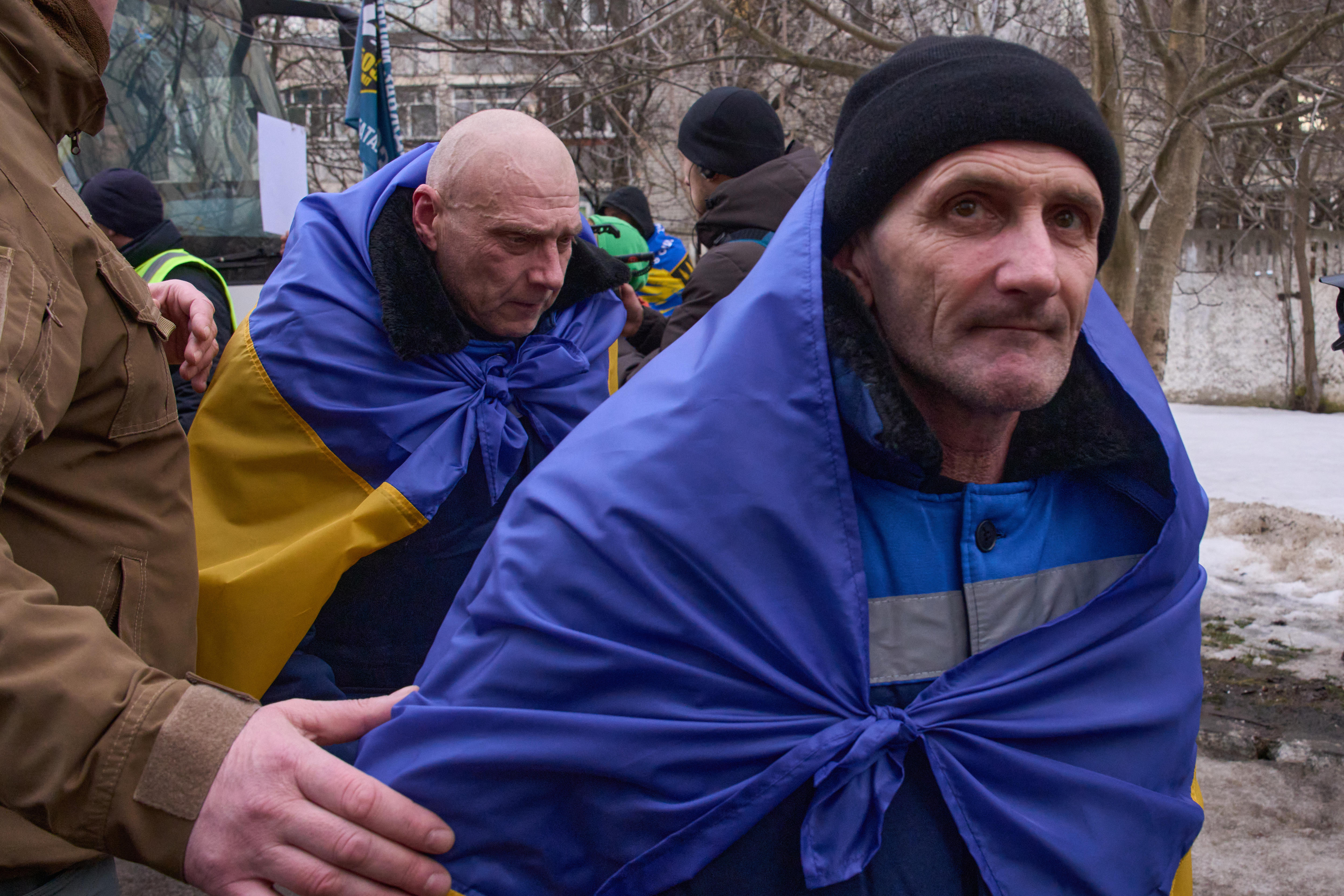 An older Ukrainian soldier, wrapped in a Ukraine flag, looks at the camera, with another soldier behind him.