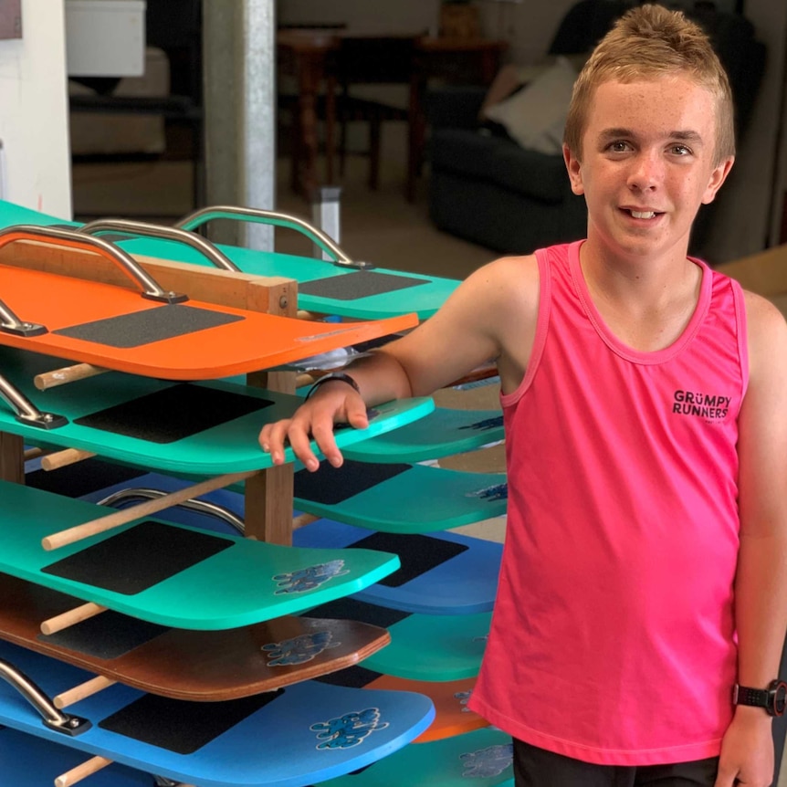Boy on right in pink singlet leaning on rack of blue, green and orange sandboards