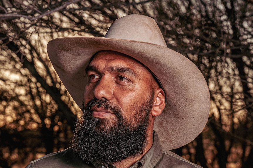 A slightly smiling Indigenous man with a beard wearing a wide-brimmed hat looks away from the camera.