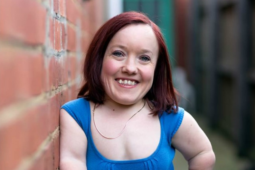 A short statured woman poses in front of a brick wall.