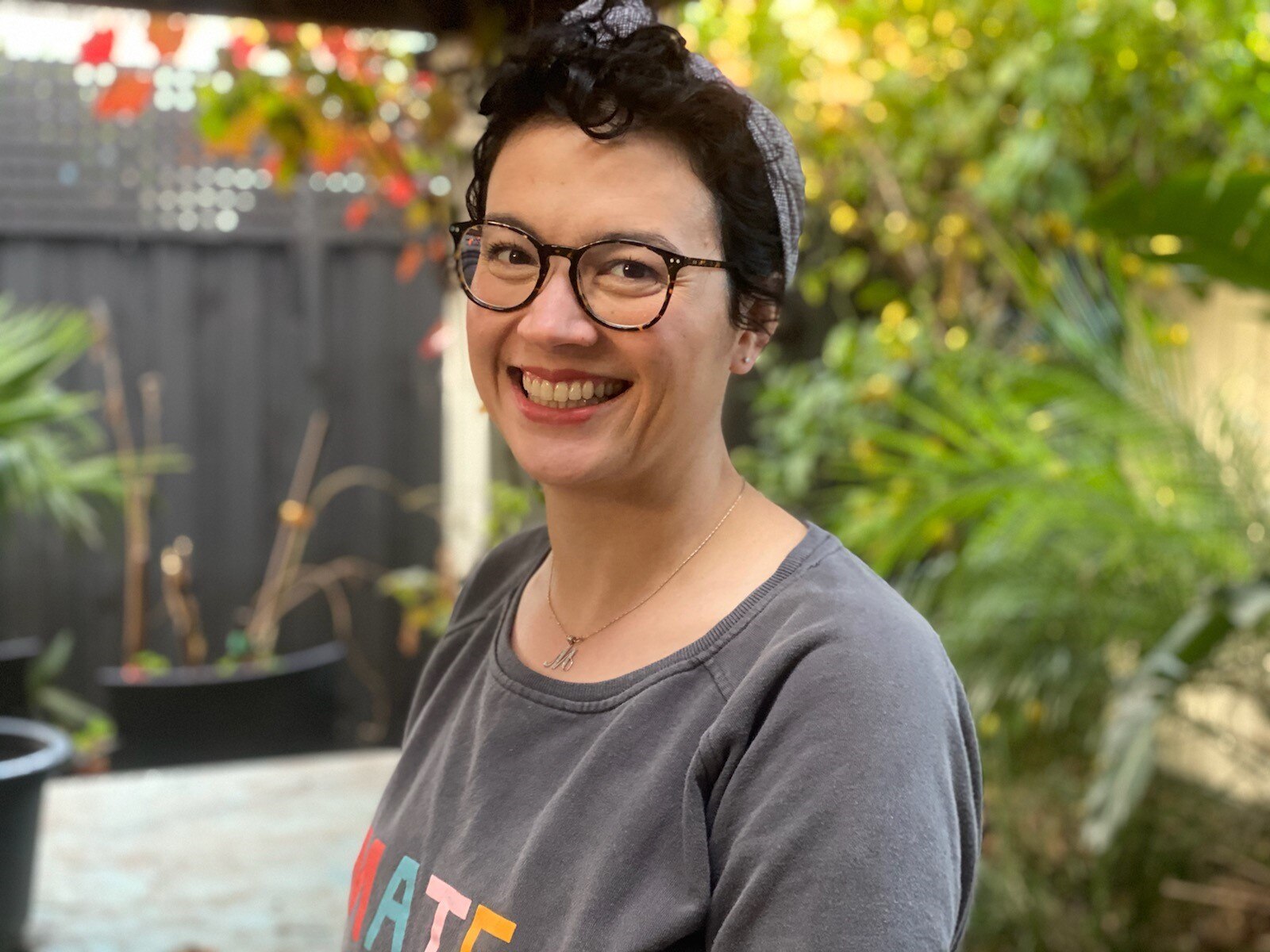 Marianne is smiling. She has short, dark hair and is wearing glasses. She is in a garden setting.