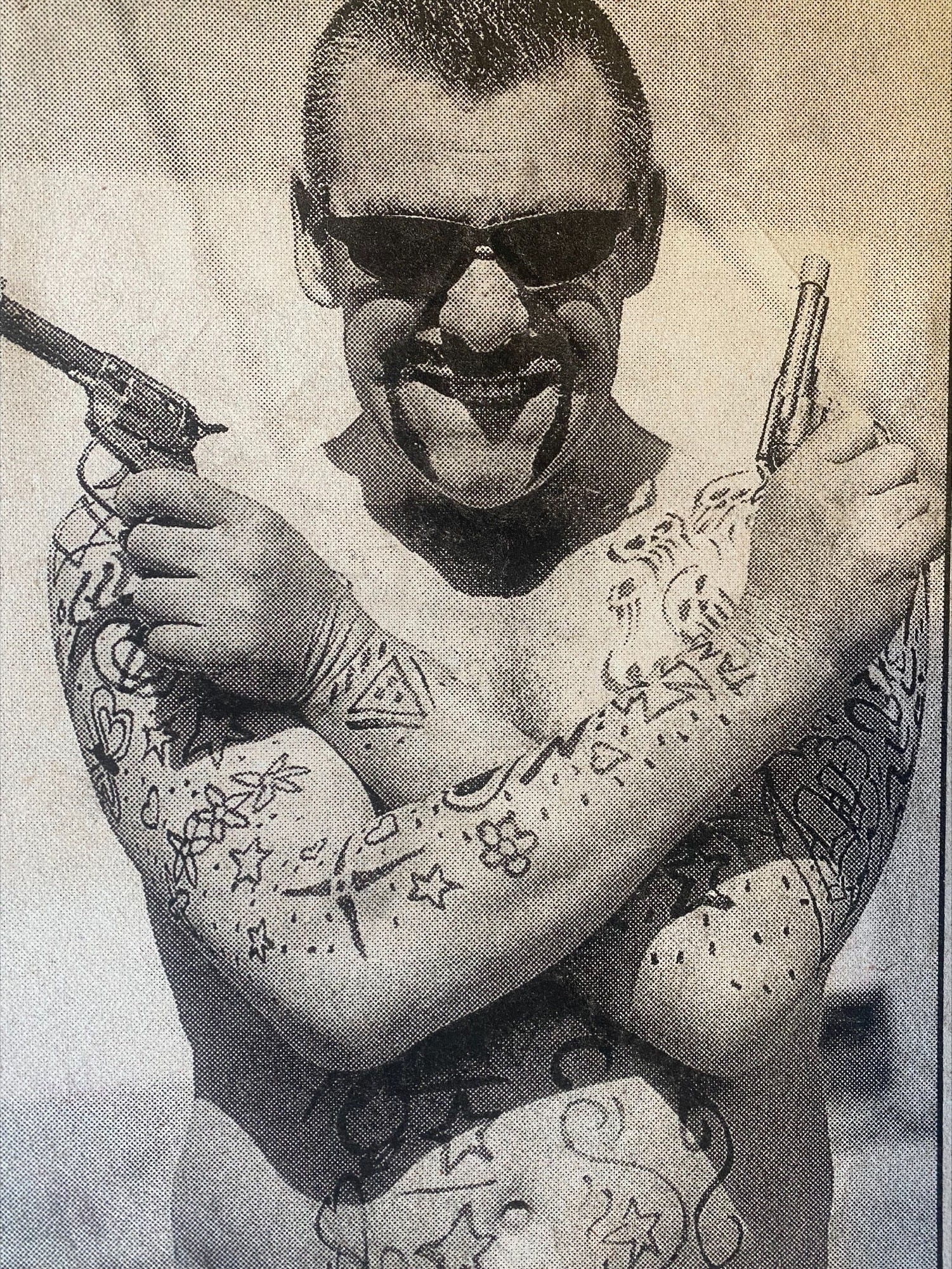 Una fotografía en blanco y negro de un hombre sin camisa con tatuajes dibujados a mano, gafas de sol y armas falsas.