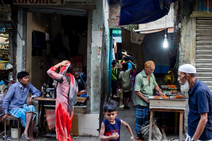 A man walks past another man making barbecue at the entrance to an alley while a woman talks to a man nearby.