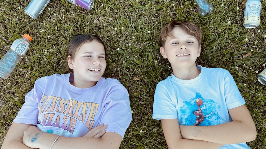 Two kids lying on grass surrounded by empty bottles and cans