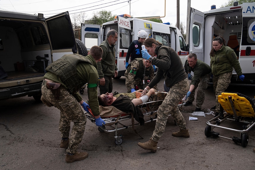 Раненый мужчина лежит на носилках, ему помогают парамедики из машин скорой помощи.