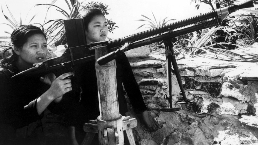 Two young North Vietnamese girls learn how to handle a machine gun which has been placed against a makeshift bamboo structure.
