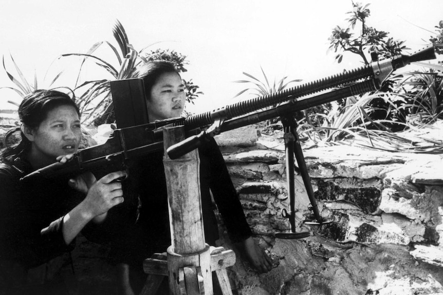 Two young North Vietnamese girls learn how to handle a machine gun which has been placed against a makeshift bamboo structure.