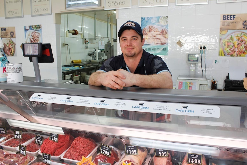 A man poses behind the counter in a butcher's shop.