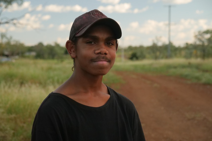 A boy with a brown cap and a black shirt stands on a dirt road