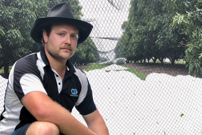 A man wearing a black, full-brimmed hat kneels beside a huge pile of hail that's fallen in an orchard.