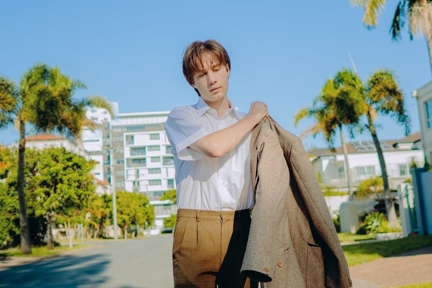 A young man stands on a tree-lined path leading to a big building, as he puts on his jacket.