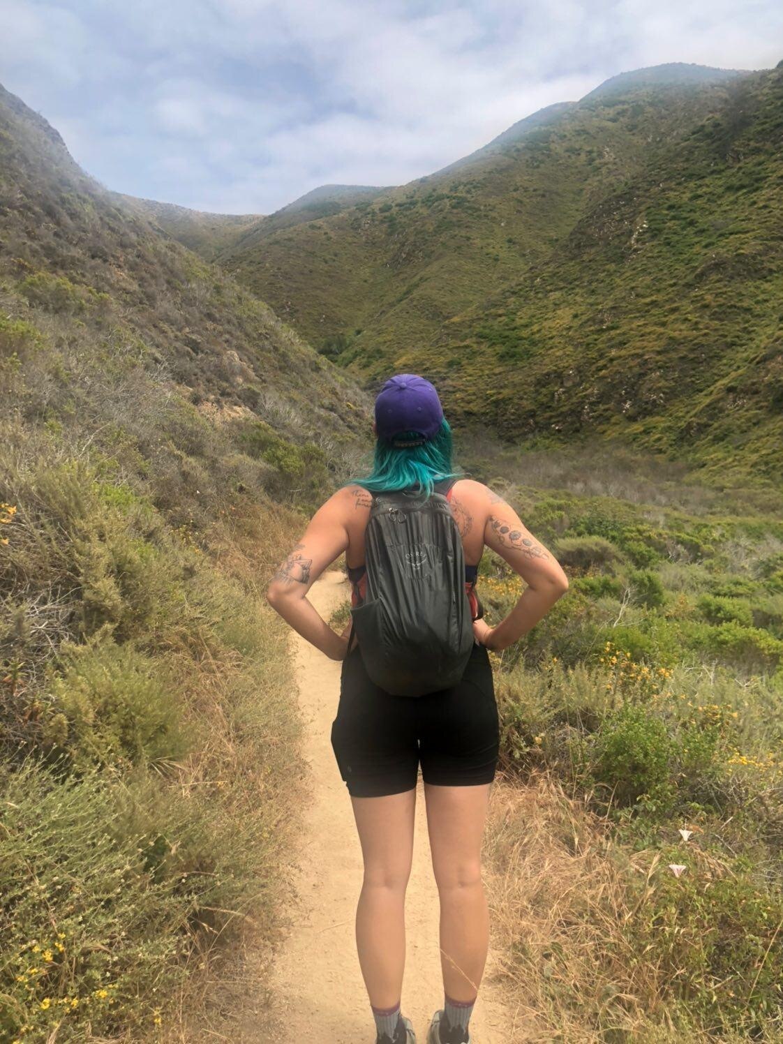 A person stands with their hands on hips, back to camera, wearing a backpack, on a trail and looking out towards mountains.