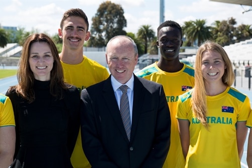 A man in a suit smiles at the camera surrounded by a group of Australian athletes