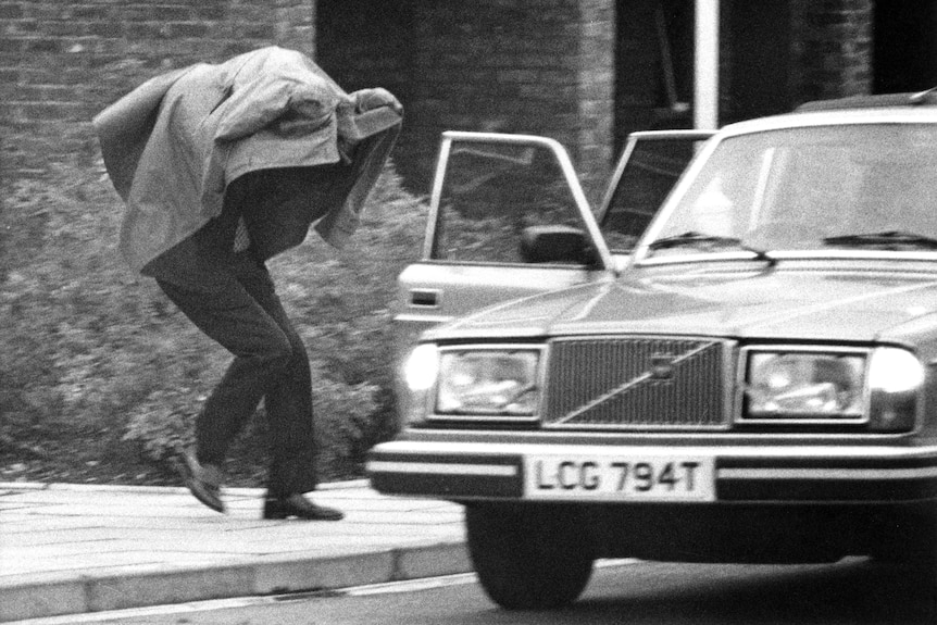 A black and white photo of a man dashing for a parked Volvo, a trench coat over his head