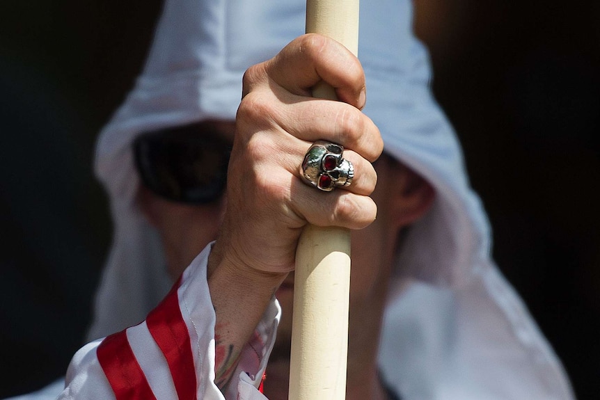 A skull ring is seen on the hand of a man wearing a Ku Klux Klan hood.
