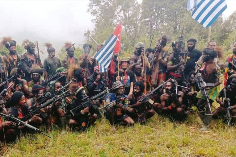 A group of West Papua rebels with many holding guns and wearing traditional headgear, New Zealand pilot holding West Papua flag.