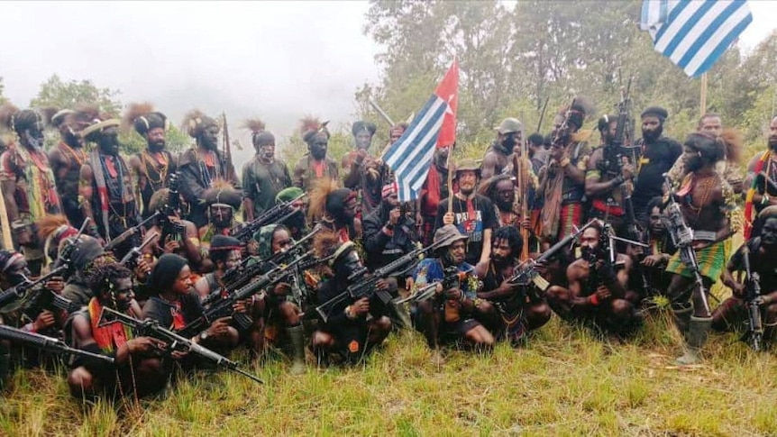 A group of West Papua rebels with many holding guns and wearing traditional headgear, New Zealand pilot holding West Papua flag.