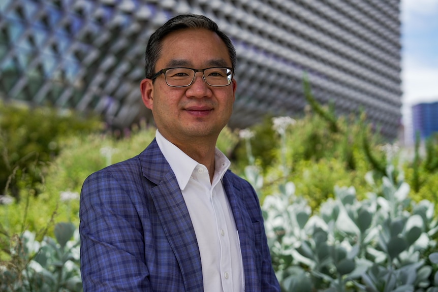 A man in a checked blue suit and glasses smiles outside a modern building.