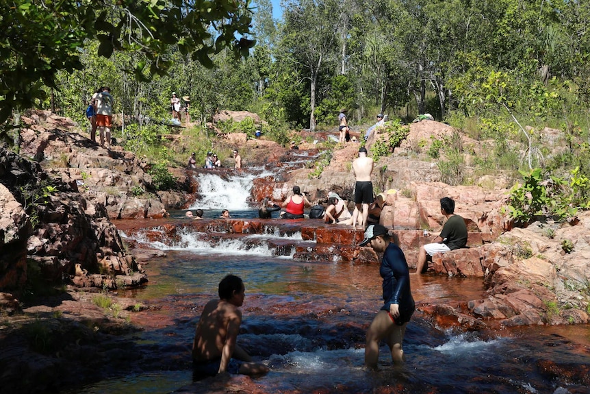 At a small waterfall, some people swim in the water while other sit on the rocks.