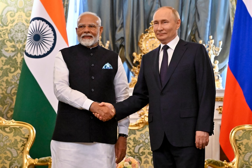 Narendra Modi and Vladimir Putin shake hands while smiling in front of an Indian and Russian flag