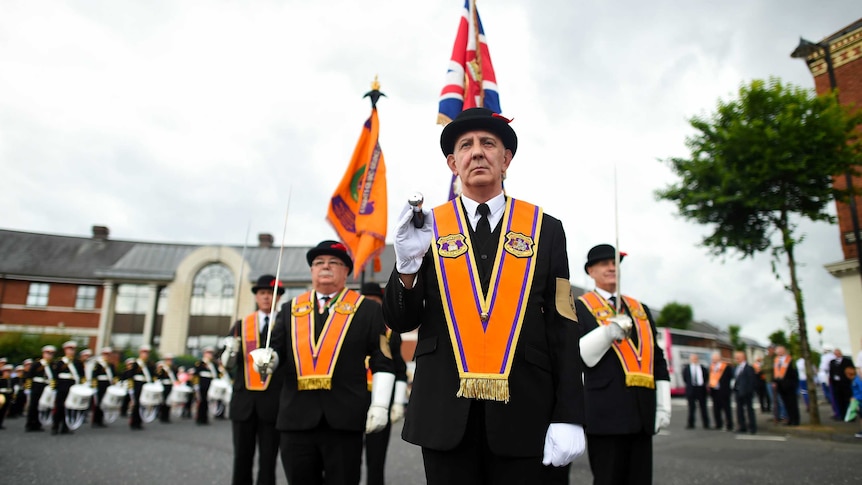 Men in bowler hats, orange collarettes and white gloves stand with flags and swords before beginning the annual Orange march.