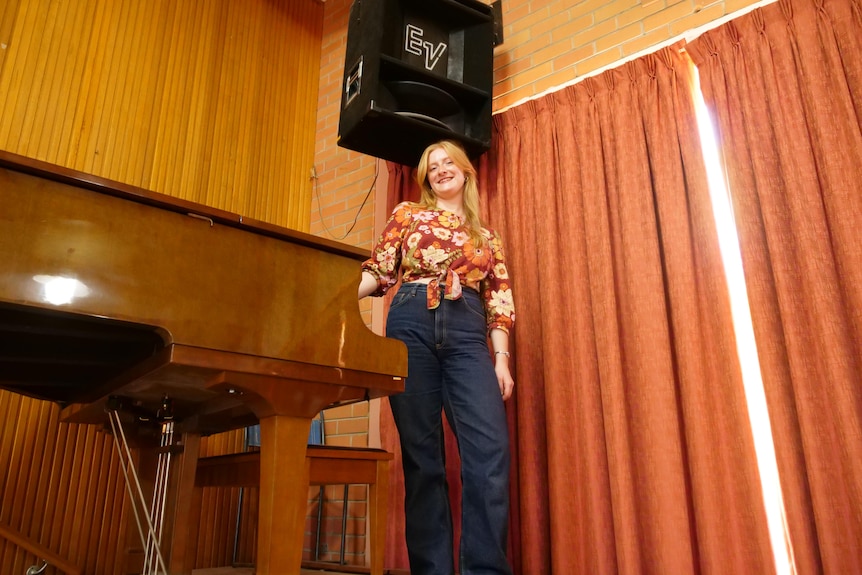 A woman wearing jeans and a floral top stands next to a piano near curtains.