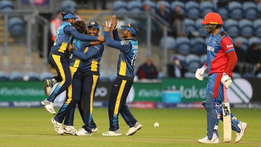 A group of cricketers jump together in jubilation as a batsman trudges off after losing the match.