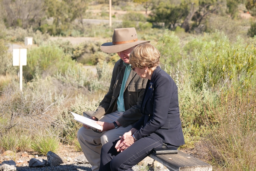 A man and a woman sit on a wood bench in bushland. 