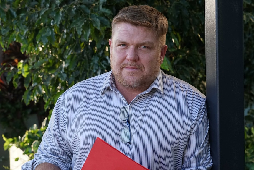 A man in a formal shirt leans on a pole and looks at the camera.