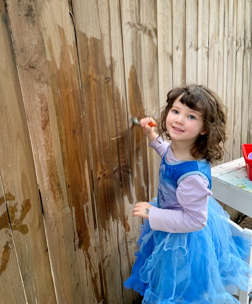 A young girl wearing a blue dress paints a fence with a paintbrush dipped in water.