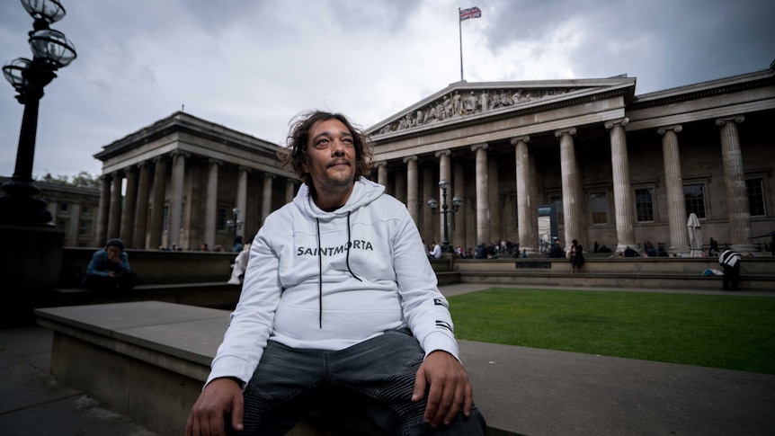 An Indigenous man in a white shirt sits outside a large, stately building.