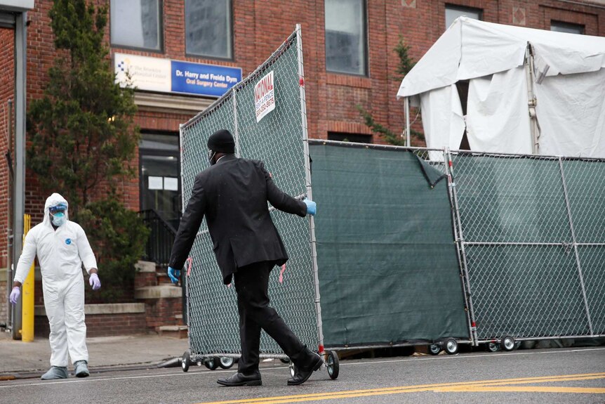 Man in a black suit pulls close a gate outside a surgery as a person in a white protective suit looks on.